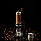 A manual coffee grinder with a dark wood handle and black metal base, shown with a small clear glass cup under the grinding head and a separate metal container on a light gray surface against a soft gray background.