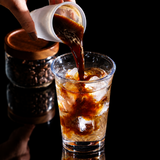 A hand pours dark coffee from a white measuring cup into a clear glass of iced coffee on a wooden table. A Binco logo is visible in the top left, and a jar of coffee beans sits blurred in the background.