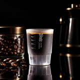 A frosted glass measuring cup with printed measurement markings and the Bingo Coffee Uppers brand logo, shown on a wooden tabletop with coffee beans and coffee equipment in a real indoor setting. A dark banner at the top reads 'Bingo Scene real shooting display'.