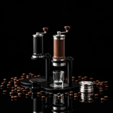 A manual coffee grinder set with a wooden-handled model and a black-handled model, both on black metal stands with clear glass cups underneath, displayed on a light gray stone surface against a textured gray wall.