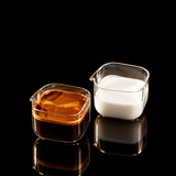 Two clear square glass measuring cups with white rims and the Binco logo, one filled with espresso and the other with milk, placed on a wooden board with coffee beans.