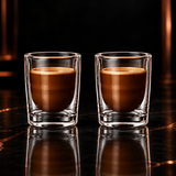 A clear double-walled glass tumbler containing a brown coffee drink, centered on a black reflective surface with a dark studio background.