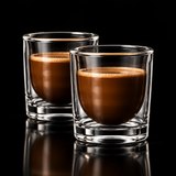 A clear double-walled glass tumbler containing a brown coffee drink, photographed against a black background with a reflective surface below.