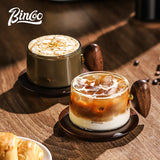 Two Binco coffee cups on a wooden table, one holding a latte with foam art and one holding an iced coffee with milk, both with dark wood handles and saucers.