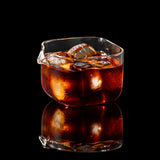 A clear glass iced coffee pitcher with a pouring spout, filled with amber coffee and ice, on a dark gray surface with coffee beans scattered nearby.