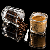 Two clear glass coffee cups with printed measurement markings, one tipped over with coffee beans spilling out and the other filled with espresso, on a white surface with dried orange slices and green leaves in the background. KEMORELA branding is visible in the top left.