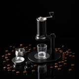 A manual coffee grinder with a matte black body and silver metal components, shown on a light gray stone tabletop with its glass carafe and small accessories nearby.