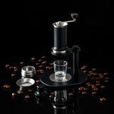 A manual coffee grinder with a matte black body and silver metal components, shown on a light gray stone tabletop with its glass carafe and small accessories nearby.