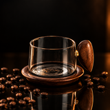 A 250ml water drop glass coffee cup in black, featuring a clear glass body, a dark brown wooden lid with a gold accent, and a matching wooden base, shown on a white surface with coffee beans scattered nearby.