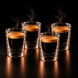 Two ribbed clear glass shot glasses filled with black coffee and topped with a layer of golden crema, shown on a dark reflective surface against a black background with steam rising.