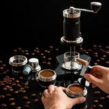 A manual coffee grinder setup shown on a wooden table with hands assembling and filling the grinder components. Large text at the top reads 'THREE TYPES OF COLLOCATION FREE CONVERSION' and labels point to capsule adapter, powder tray, and powder bowl.