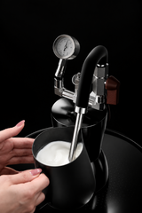 A close-up of a milk steaming apparatus with a curved metal steam wand and gauge, shown dispensing steam into a cup of milk. A person's hands are visible holding the cup, and the scene includes promotional text about storing steam in a drier.